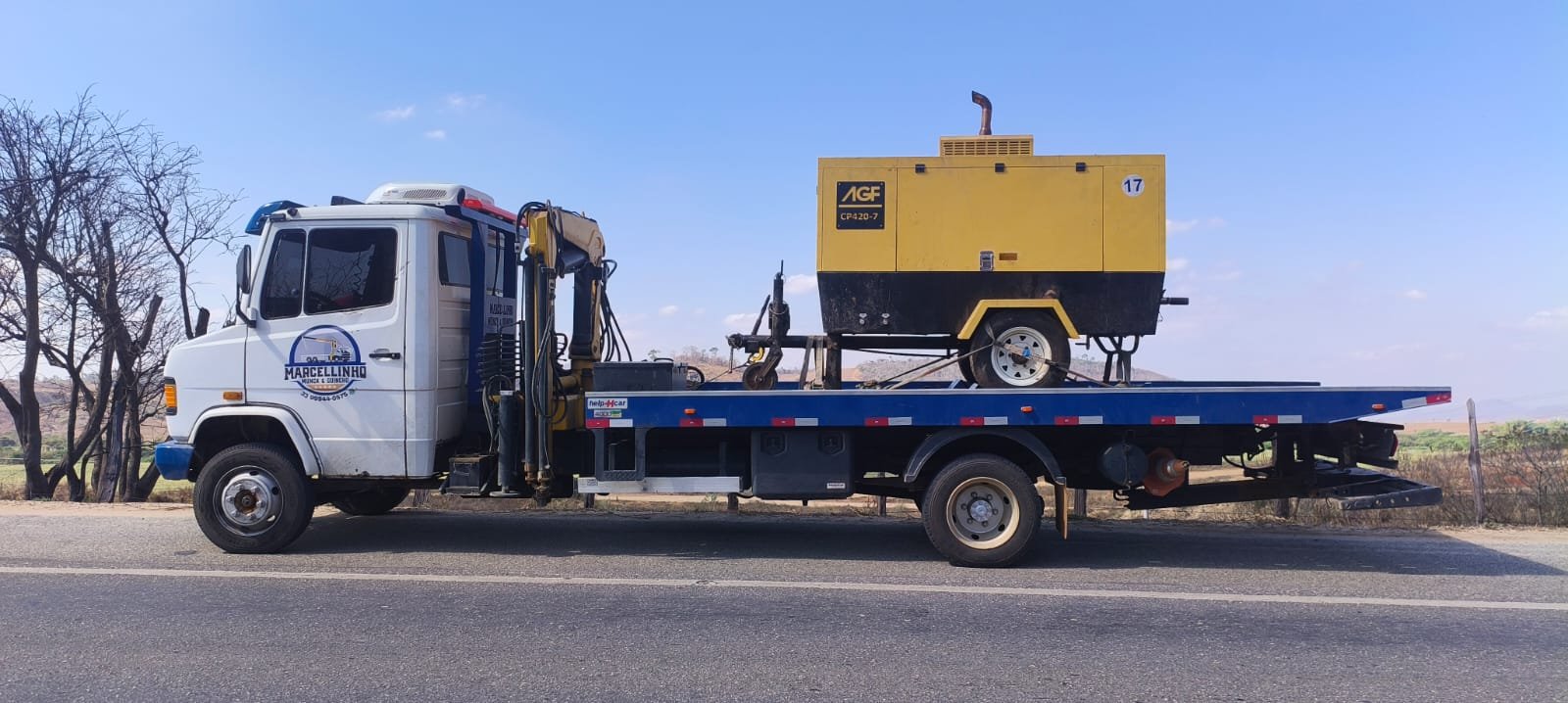 Caminhão branco com plataforma azul e munck, transportando um gerador amarelo