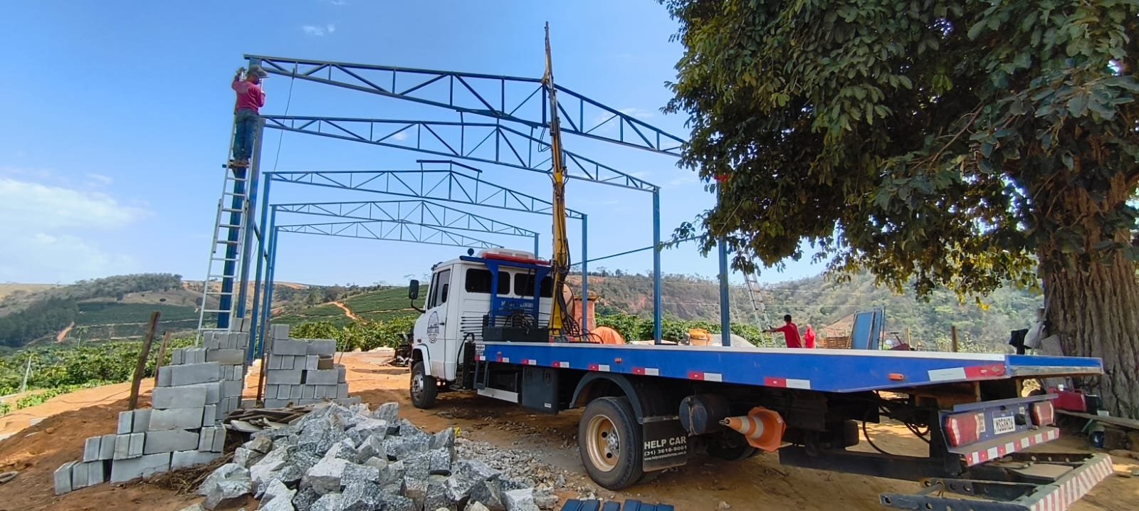 Munck levantando uma estrutura metálica em uma obra rural, com o caminhão ao lado.