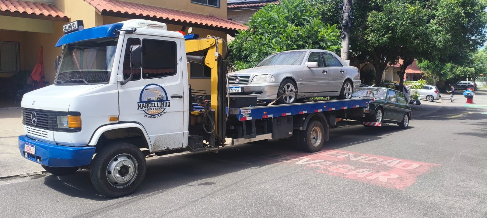 Caminhão estacionado na rua com um Mercedes prata na plataforma e uma Brasília verde engatada.