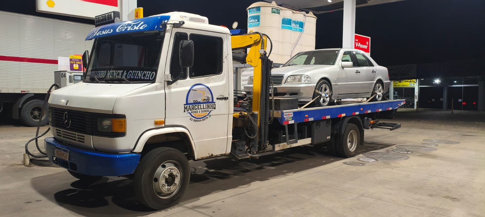 Caminhão em um posto à noite com um Mercedes prata preso na plataforma.